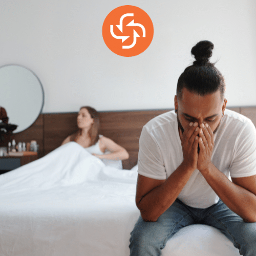 Man sitting on the edge of the bed looking stressed, while a woman sits in the background, symbolizing relationship or sexual health issues.