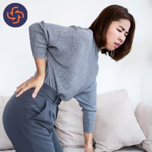 Woman standing and holding her lower back with a pained expression, showing signs of back pain or discomfort.