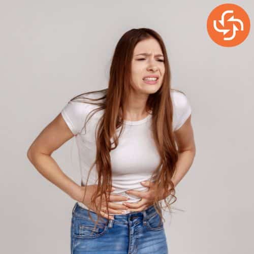 Woman in a white shirt clutching her stomach with a pained expression, indicating abdominal pain or cramps.