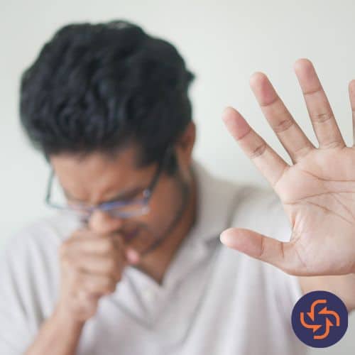 Man coughing into his fist while raising his other hand forward, showing discomfort or illness.