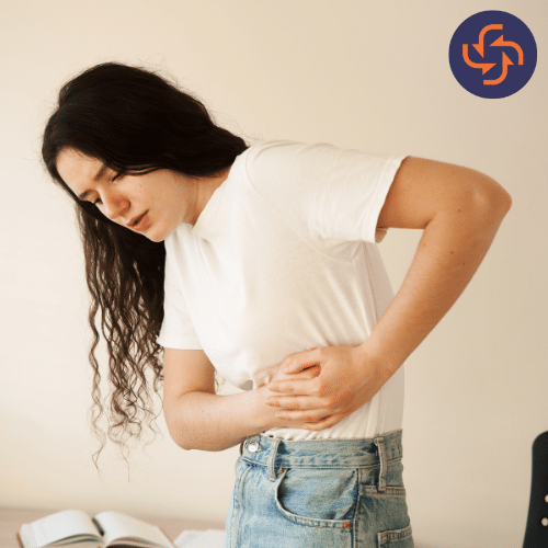 Young woman in a white t-shirt and jeans bending slightly while holding her side in pain, indicating abdominal or liver discomfort.
