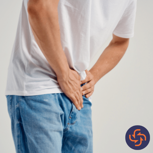 Close-up of a man in jeans and a white t-shirt holding his lower abdomen and groin area, indicating pain or discomfort.