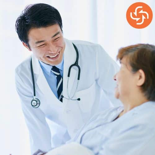 Male doctor in a white coat with a stethoscope smiling while talking to a patient in a hospital bed.