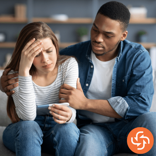 Couple sitting together, with the woman holding her head in worry while looking at a pregnancy test, as her partner supports her.