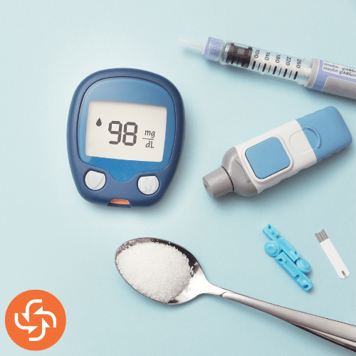 Flat lay of a glucometer, insulin syringe, test strips, and a spoon of sugar on a blue background, representing diabetes management and blood sugar monitoring.