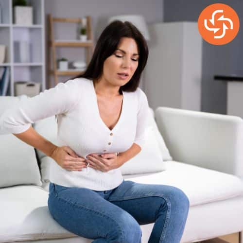 Woman sitting on a couch holding her abdomen with a pained expression, indicating stomach pain or cramps.