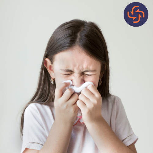 Little girl in a white t-shirt sneezing into a tissue with eyes closed, representing flu, cold, or allergy symptoms.
