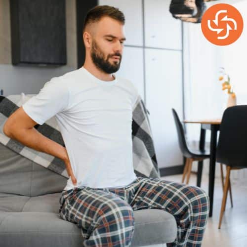 Man sitting on a couch in pajamas, holding his lower back with a pained expression, indicating back pain or discomfort.