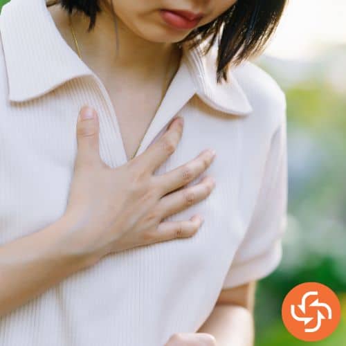 Woman in a light shirt holding her chest with one hand, appearing to experience chest discomfort or pain.