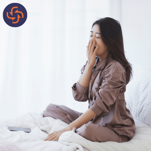 Young woman in pajamas sitting on bed with a hand covering her mouth while yawning, indicating tiredness or sleepiness.