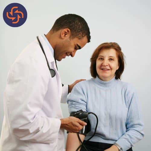 A smiling male doctor in a white lab coat with a stethoscope around his neck checking the blood pressure of a female patient.