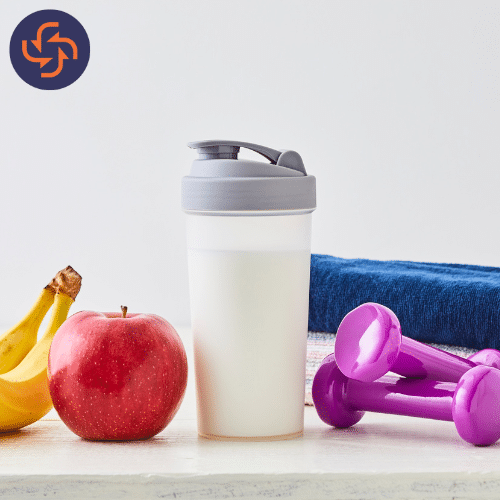 Protein shake bottle placed with an apple, banana, dumbbells, and a towel, representing fitness, nutrition, and healthy living.