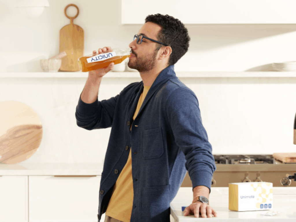 A man with glasses and a blue cardigan drinking an amber-colored liquid from a Unicity glass bottle in a kitchen, with a box of Unimate supplement visible on the counter in the foreground.